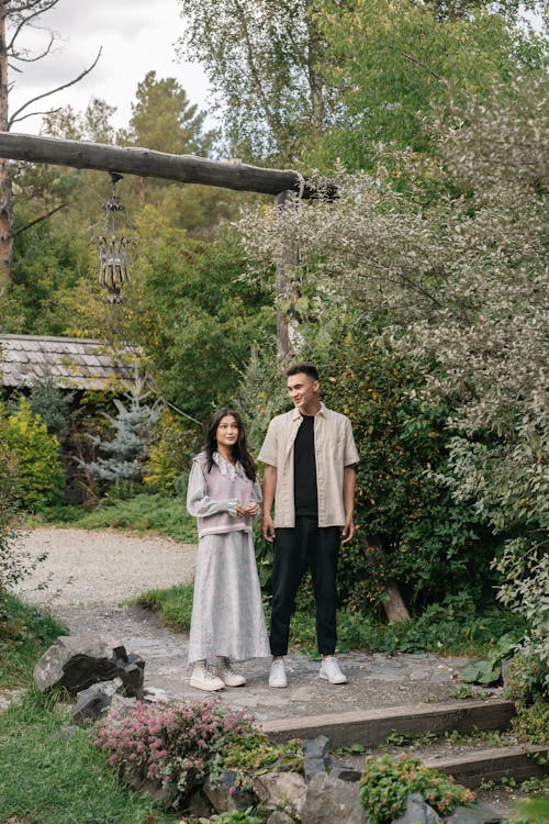 a young asian couple stands smiling on a path in a lush green garden.  the man is wearing a light beige shirt and dark pants, while the woman wears a long, light gray dress.  they are positioned in front of a rustic wooden archway, with various plants and trees surrounding them.  the setting is serene and peaceful, suggesting a romantic atmosphere.  the overall feel is one of calm and happiness.