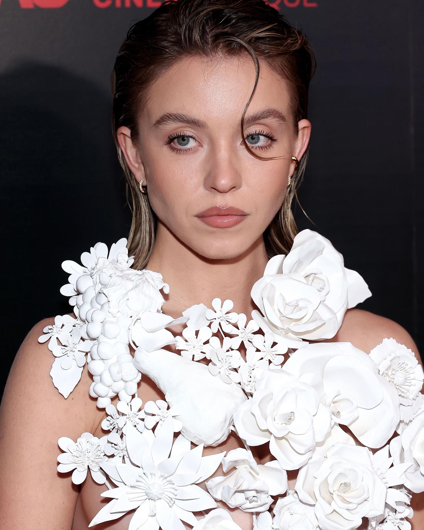 loseup portrait of a young woman with wet, styled hair, wearing a stunning white dress adorned with large, sculpted white flowers. the dress resembles a floral arrangement.  her makeup is subtle and elegant. she is looking directly at the camera. the background is dark and out of focus, suggesting a red carpet event or formal setting.