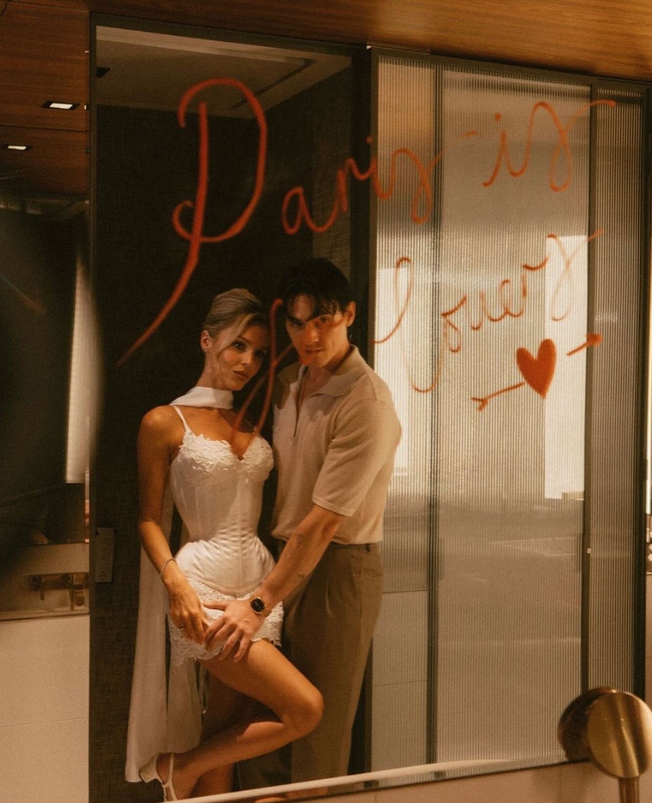 a couple is standing in front of a mirror, holding each other, looking at each other lovingly.  the words "paris" and "forever" are written in red on the mirror, and there is a heart and arrow drawn on the mirror.  the woman is wearing a white dress and the man is wearing a light beige polo shirt.  the couple looks very much in love, as if they are about to share a kiss.   the romantic ambiance is created by the lighting of the bathroom and the love message written on the mirror.  the setting is a beautiful parisian bathroom, adding to the romantic setting.