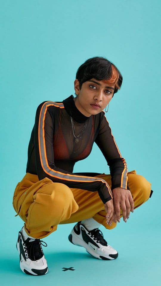 a young woman in black mesh shirt with yellow and orange stripes and yellow pants is sitting with her knees bent and feet together in a crouched position. she's wearing black and white sneakers. she is looking at the camera. the background is a light blue color.
