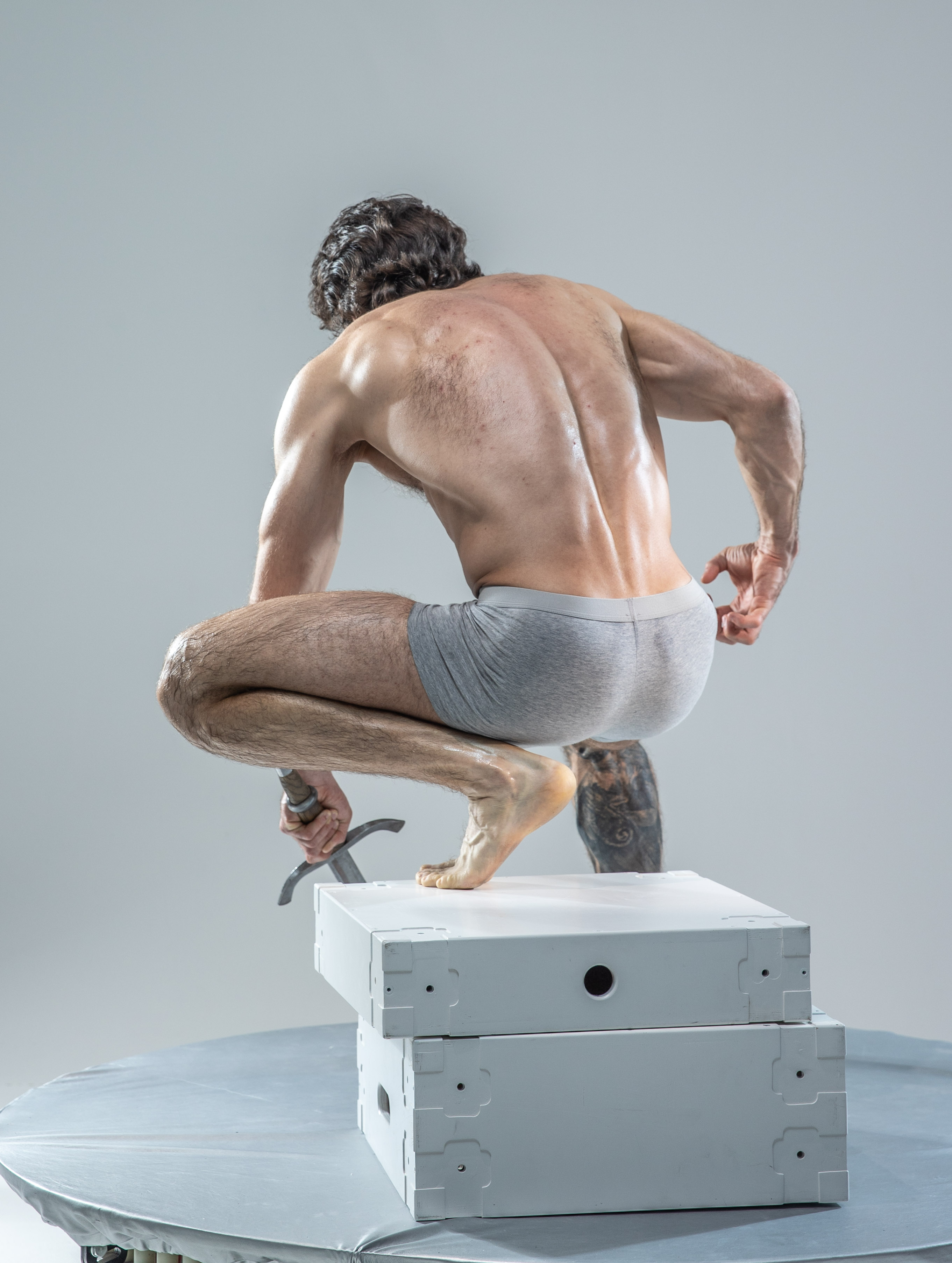 a shirtless muscular man with short, dark, curly hair is captured from a rear view, crouching on two white boxes in a photography studio setting. he holds a sword in his right hand, the point of which is partially obscured by the box. his left hand is clenched in a fist. the man's physique and the dynamic pose suggest action and power. the studio environment provides a neutral backdrop, focusing attention on the man's form and the sword he wields.