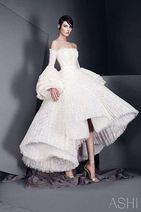 a woman is posing in a white lace highlow dress with large, voluminous sleeves.  the dress is fitted at the bodice and then flows out into a  full skirt with a highlow hemline. the woman is wearing her hair pulled back and is looking at the camera with a serious expression.  she is standing on a grey surface with the word "ashi" written in the bottom right corner of the image.  the dress is part of a fashion photoshoot.