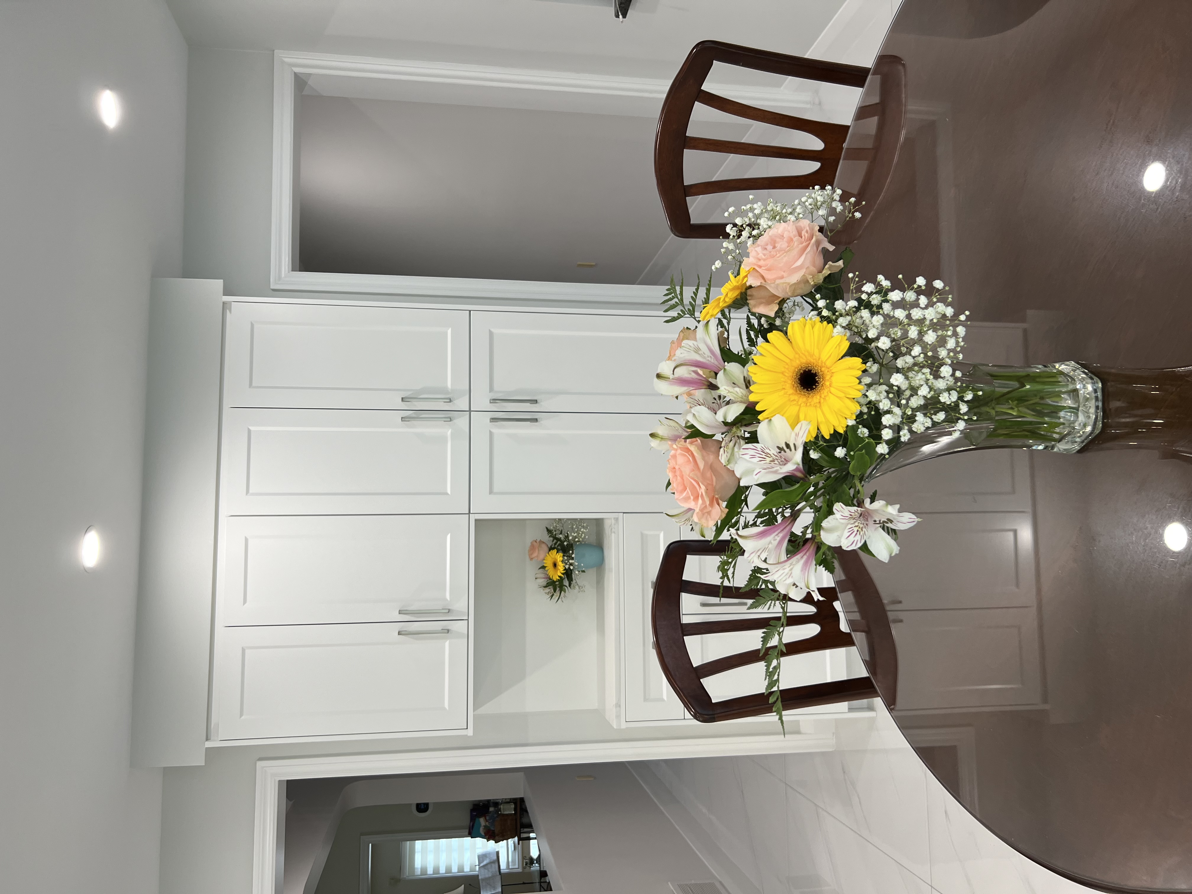 the image shows a bright and modern kitchen with newly installed white builtin cabinets.  a dark brown round dining table sits in front of the cabinets, with two dark wood chairs partially visible. a beautiful bouquet of flowers in a clear glass vase is the centerpiece on the table. the overall feel is clean, elegant, and contemporary.  the flooring appears to be a lightcolored tile or marble. the background shows a hallway and a glimpse of another room.  recessed lighting is visible in the ceiling.