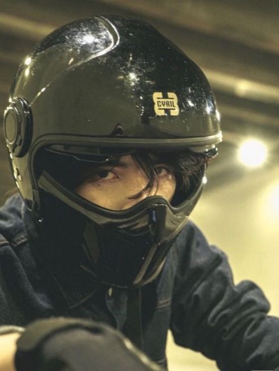 the image shows a closeup of a person wearing a black cvril fullface motorcycle helmet. the helmet is shiny and has a somewhat futuristic look.  the person is wearing a dark denim jacket and their face is partially obscured by the helmet's face mask. the background is blurry and dark, creating a moody atmosphere. the overall style is edgy and urban.  the focus is on the helmet and the person's determined or serious expression.