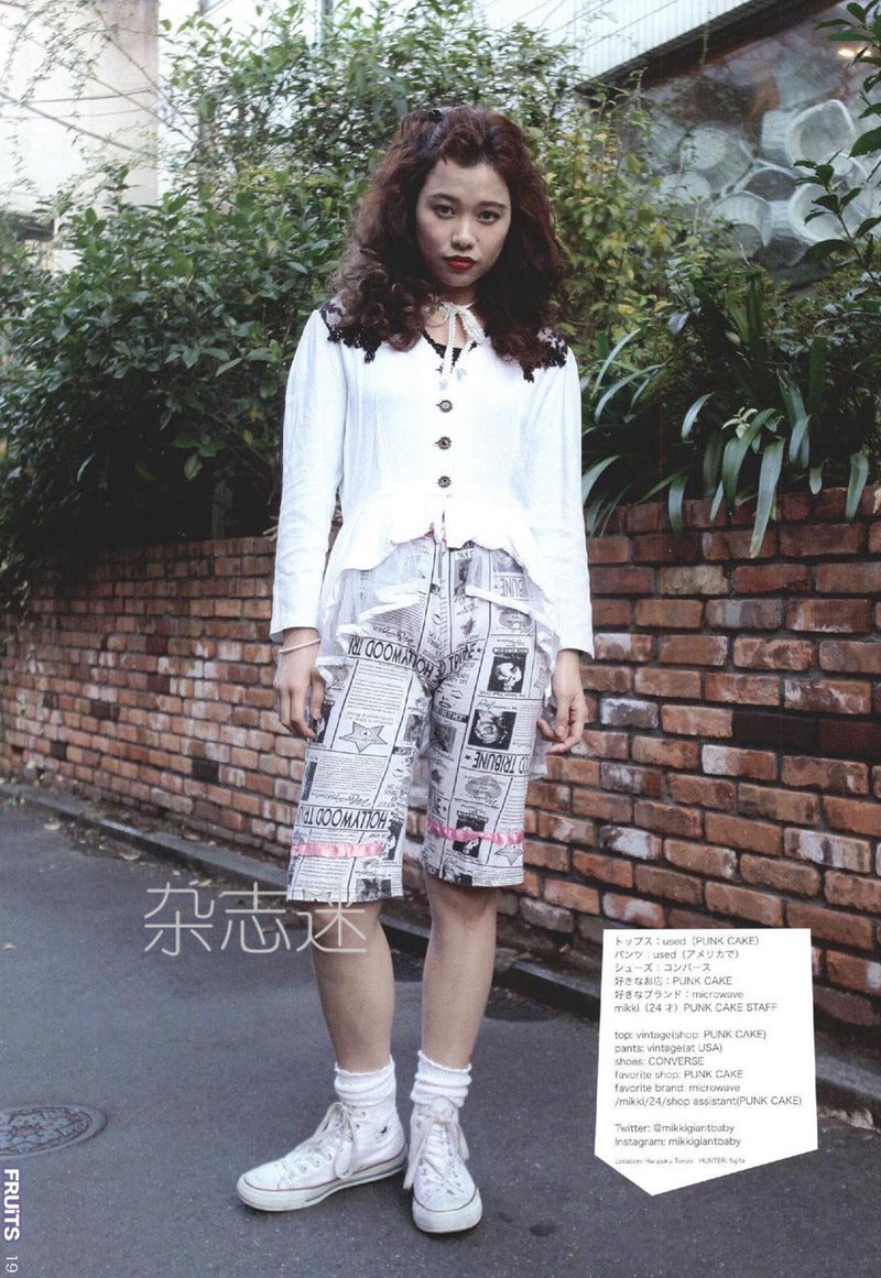 a young woman with curly hair is standing on a sidewalk in front of a brick wall.  she is wearing a white top, printed shorts, and white sneakers. the image appears to be taken for a japanese street fashion magazine, as indicated by the japanese characters on the bottom right corner.