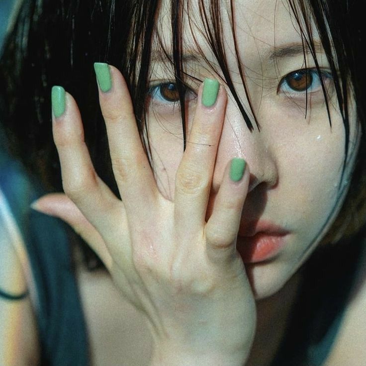 a young asian woman with green nail polish on her fingers. she is looking down at the camera and has her hand covering her face. her hair is parted in the middle and she is wearing a black top.