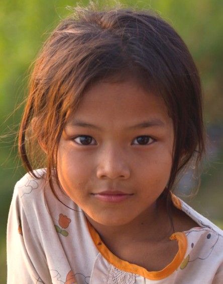 a closeup portrait of a young girl with brown hair and brown eyes. she is wearing a lightcolored shirt with a yellow trim and a pattern of flowers. the girl is looking directly at the camera with a neutral expression. the background is blurred and green, suggesting an outdoor setting.