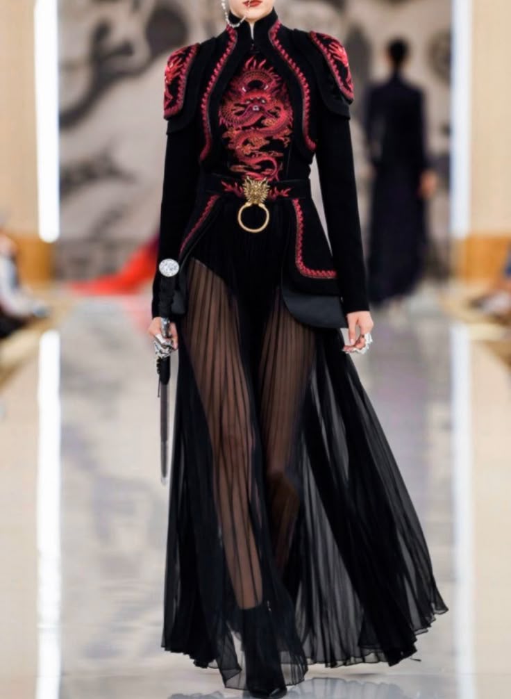 a model walks the runway in a highfashion black dress with red dragon embroidery on the bodice, a high collar, and long sleeves. the dress features a black bodysuit with a sheer pleated skirt overlay, and a belt with a distinctive dragon buckle. details include shoulder pads and tassels, and the model is wearing a large ring. the background shows other models and the runway setting.