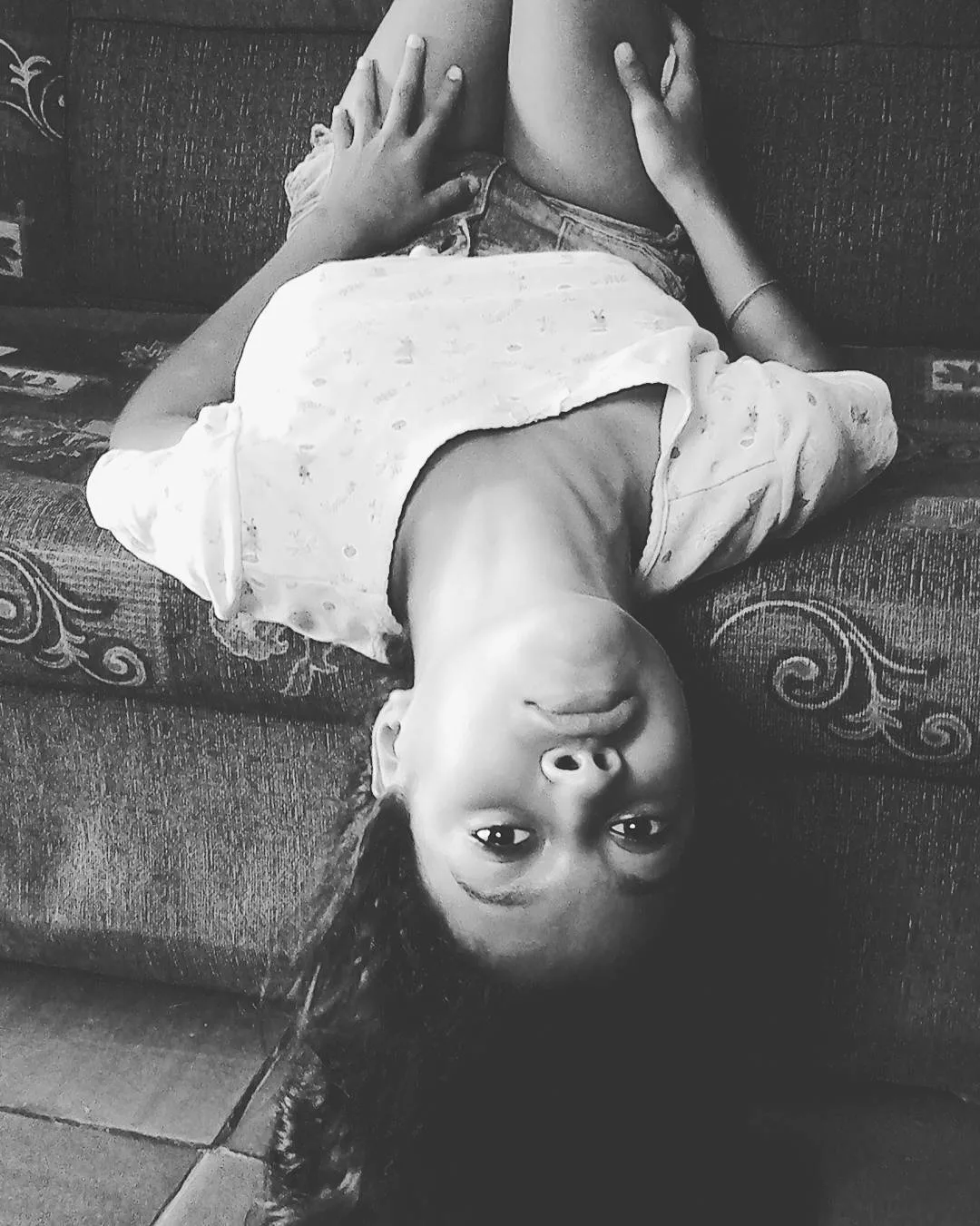 a woman in a tshirt and shorts is laying upside down on a couch. the image is in black and white. she is facing the camera, and has one hand on her leg.