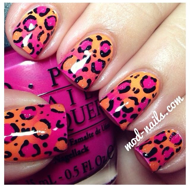 a closeup image of a woman's hand with leopard print nail art. the nails are painted in a bright pink and orange color, with black leopard spots. the image is taken at a closeup angle so that the details of the nail art are visible. there is also a bottle of nail polish on the left side of the image. the background is blurry.