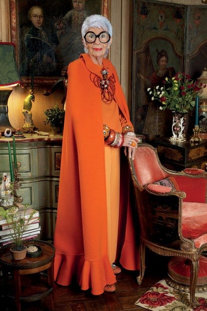 the image is a fulllength portrait of iris apfel, a renowned fashion icon. she is standing in what seems to be her living room, which is richly decorated with antique furniture and art. apfel is wearing a striking, floorlength orange cape with a ruffled hem and an orange dress underneath. she accessorizes with large statement jewelry including many bracelets and a bold necklace. her style is bold, eclectic, and undeniably unique.  the background showcases her home's sophisticated interior design and a selection of artwork, reinforcing her image as a person of refined taste.  the overall mood of the photograph is one of elegance and personality.