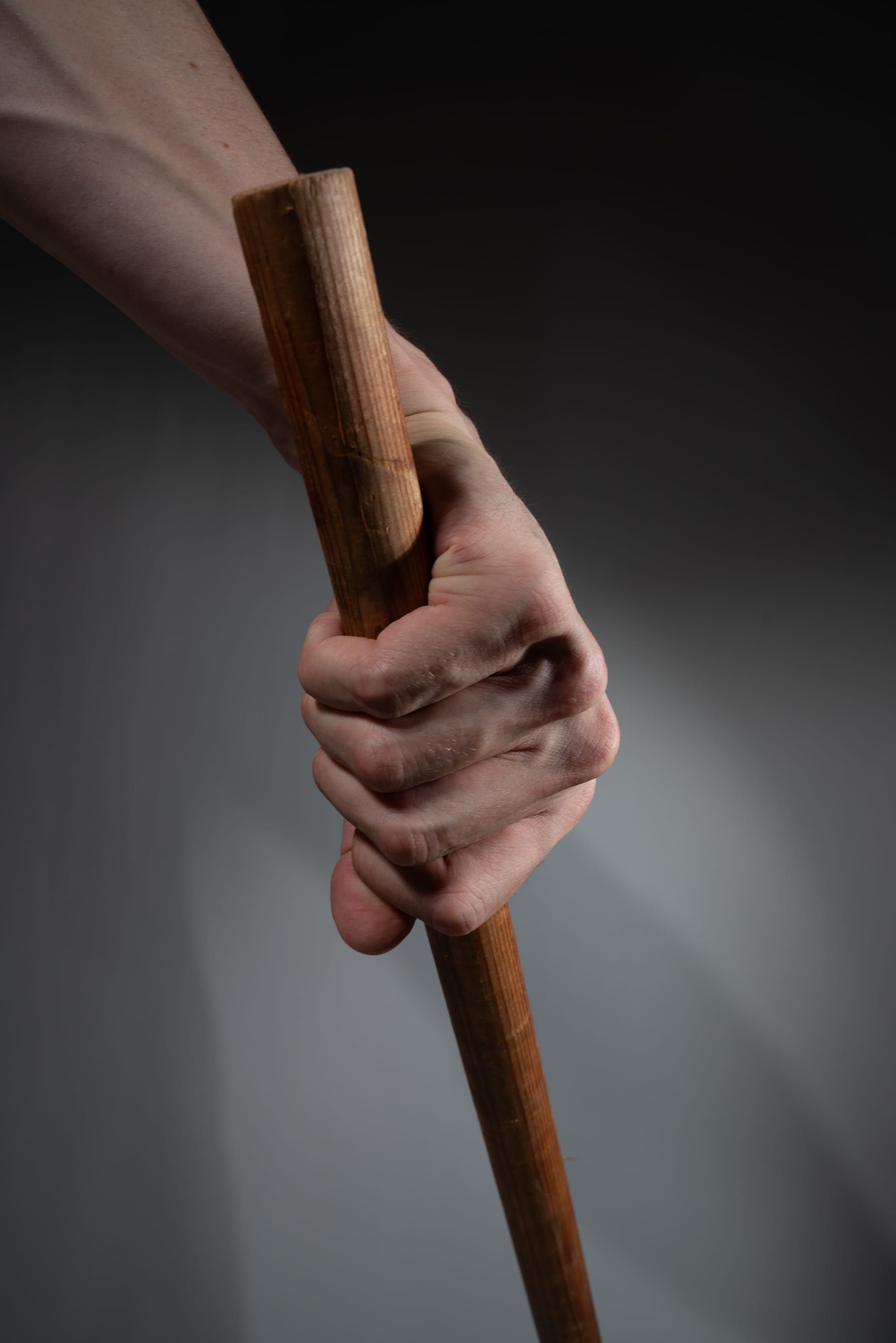 a closeup shot of a man's hand tightly gripping a wooden stick against a gray background. the hand is illuminated with studio lighting, creating strong shadows that highlight the tension and strength of the grip.