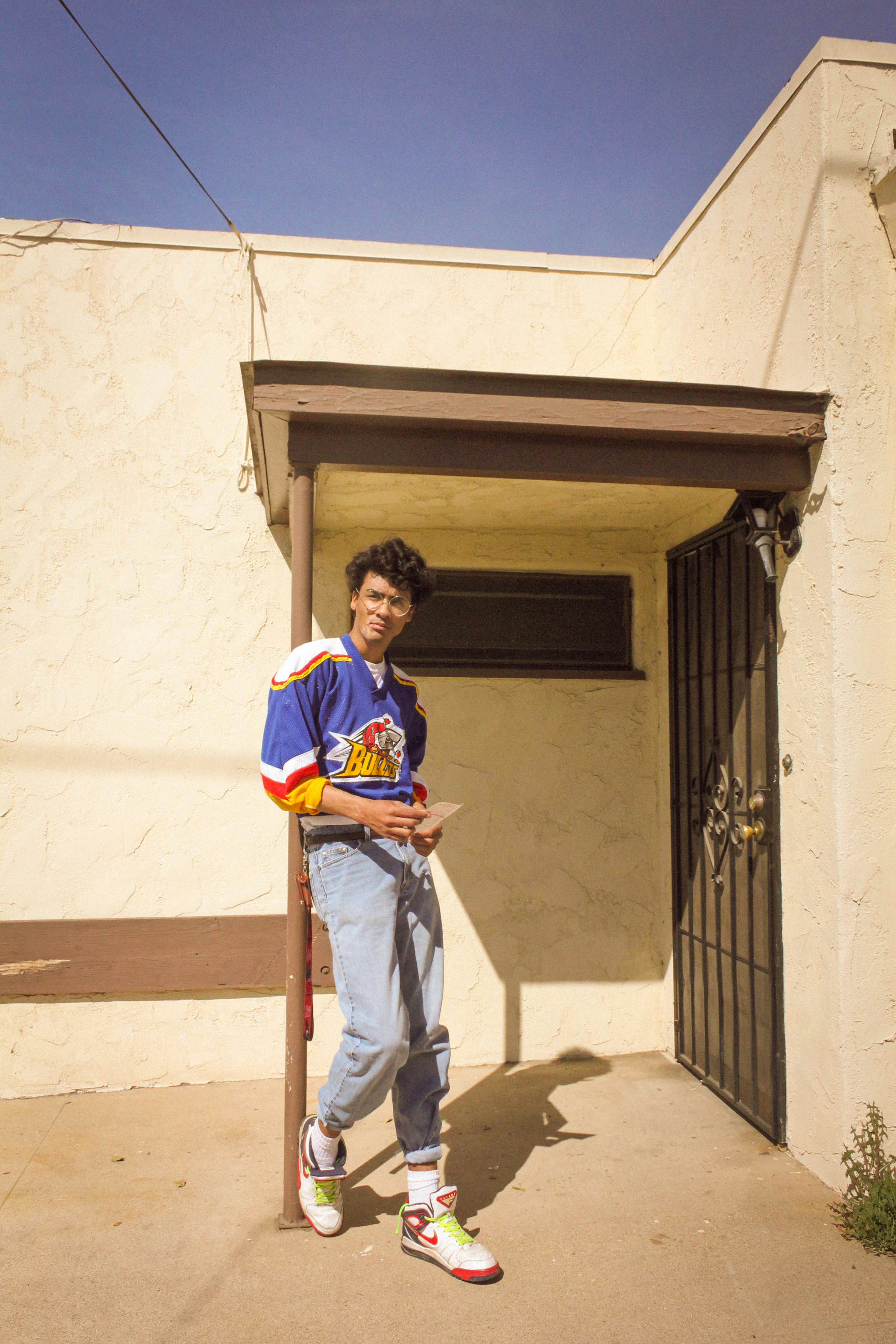 a young man, possibly a teenager, stands casually on the porch of a light beige stucco house. he's dressed in 1990sstyle clothing, including a bright blue, yellow, and red hockey jersey with a cartoonish logo, lightwash denim jeans, and hightop air jordan sneakers in a white, red and neon green colorway. he holds a piece of paper or letter in his hands. the photo is taken outdoors in bright sunlight. the background features a simple house exterior and a metal security gate.  the overall aesthetic suggests a casual, relaxed mood, reminiscent of 1990s style and culture. the photo appears to be taken with film, giving it a vintage quality.