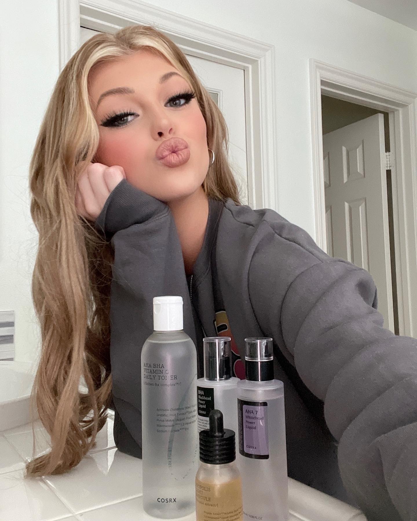 the image shows lorén gray in a selfie with skincare products on a bathroom counter. she has blonde hair and is wearing a grey sweatshirt. she is puckering her lips. the skincare products are from cosrx, including aha bha vitamin c daily toner, aha 7 whitehead power liquid, and bha blackhead power liquid.