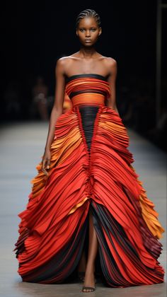 a black woman model is walking down the runway in a red and orange dress with black accents. the dress is very flowing and has a lot of layers. the model is wearing minimal makeup and has her hair pulled back. she is looking down at the ground. the dress is a very dramatic and eyecatching piece. the model is wearing a beautiful red and orange dress that is layered and flows gracefully. the dress has a black bodice and black accents. she is posing for the camera and looks stunning.