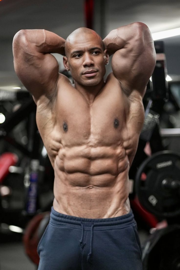 a muscular man stands with his arms crossed behind his head, showcasing his ripped physique. he has a defined sixpack and prominent muscles in his arms, chest, and shoulders. the background of the image suggests a gym setting with weights and equipment. the man appears confident and determined, highlighting the dedication and discipline involved in achieving such a level of fitness. this image represents the pursuit of physical excellence and the dedication required to build a strong and sculpted body.