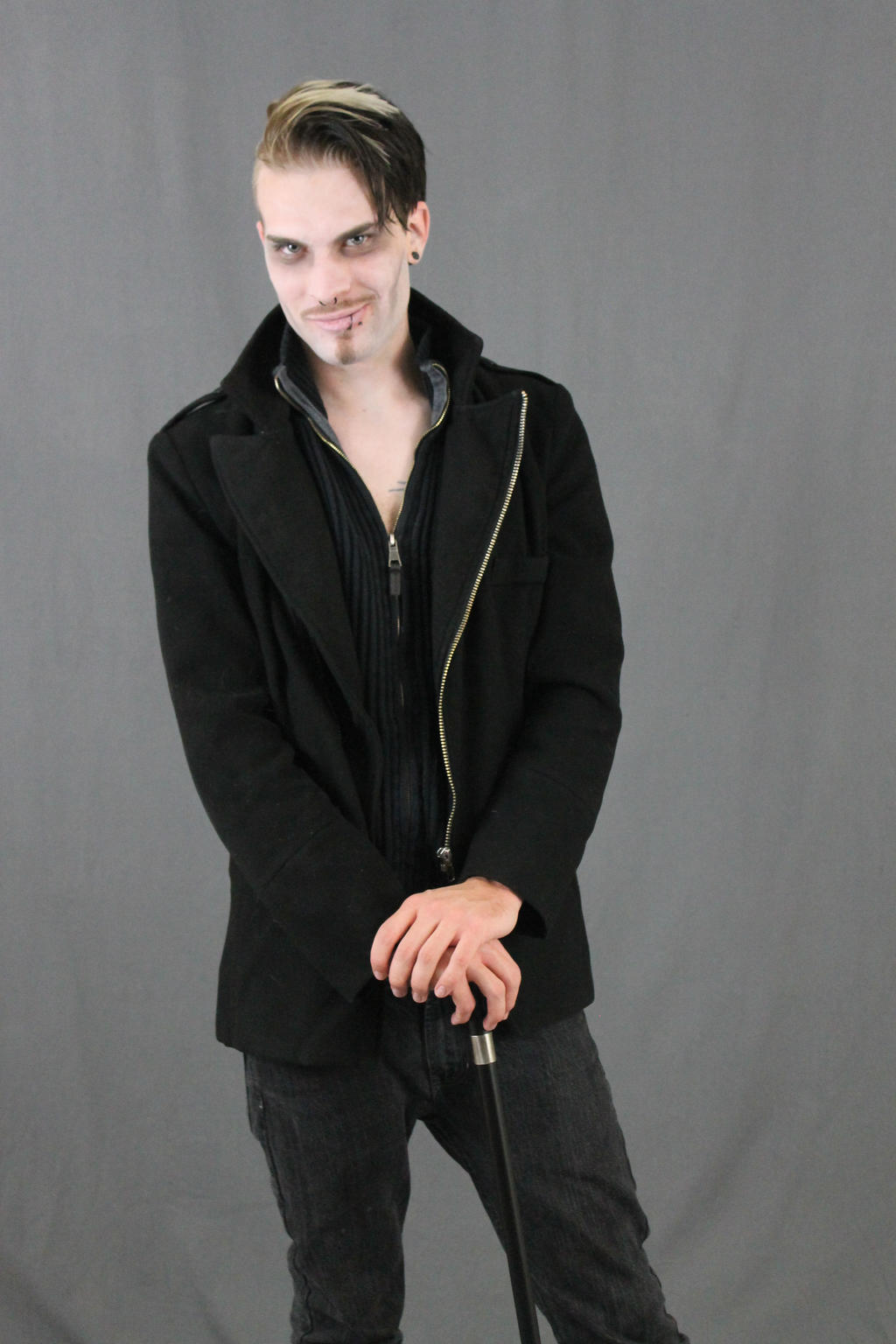 a young man with a pale complexion and blonde hair styled into a side part is standing against a grey background. he's wearing a black coat, black jeans, and is holding a black cane. his makeup is subtle, giving him a slightly gothic or alternative look. he has a smirk on his face, appearing confident and stylish. the overall image is sharp and welllit, with a focus on the model and his attire.