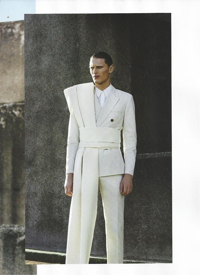 a man in a white suit with a wrap around his waist and shawl stands in front of a stone wall.