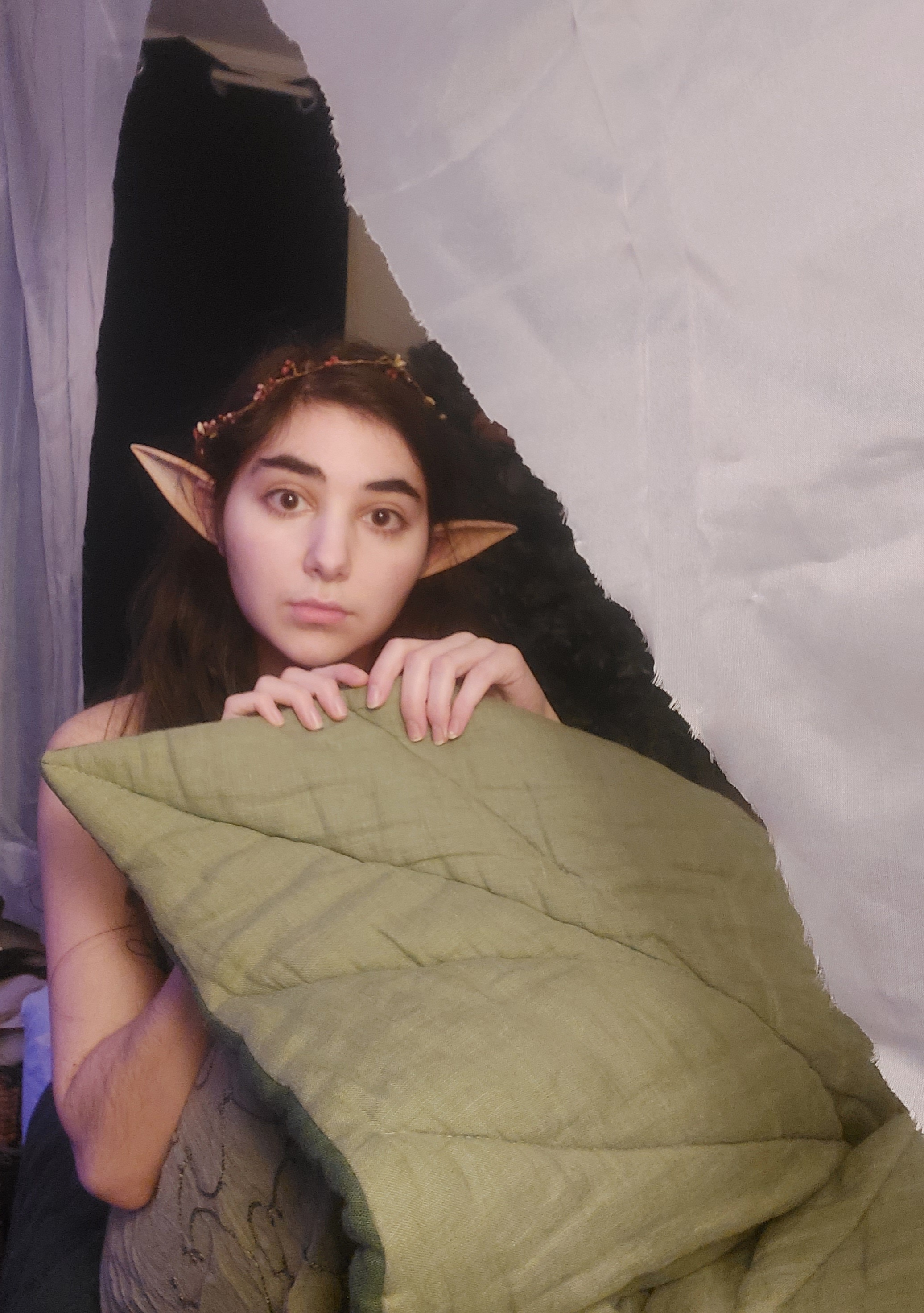 loseup portrait of a young woman cosplaying as an elf. she is wearing elf ears and a leafshaped headband. she is partially hidden behind a green pillow and a white sheet.  the setting appears to be indoors, possibly a makeshift tent or fort.  the woman's expression is somewhat shy or pensive.
