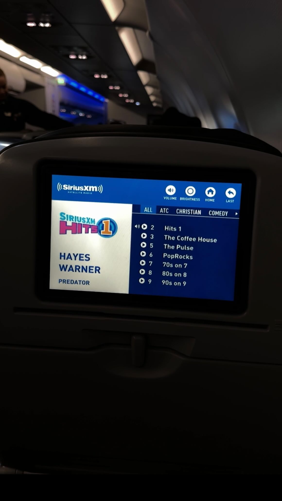 the image shows an airplane seatback screen displaying the siriusxm music channels selection. the channel "hits 1" is selected, with the song "predator" by hayes warner playing. various other music channels like "the coffee house", "the pulse", "poprocks", "70s on 7", "80s on 8", and "90s on 9" are also listed. the screen is part of the inflight entertainment system on an airplane. the airplane's ceiling is visible in the background.