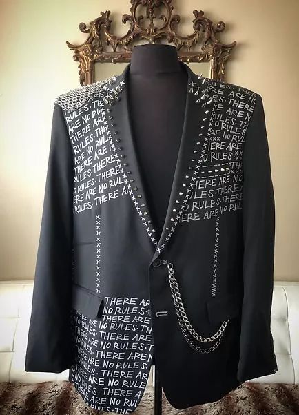 a close up shot of a black men's blazer with the phrase "there are no rules" repeated on the fabric with studded accents and a chain.  it is styled in a punk rock fashion.