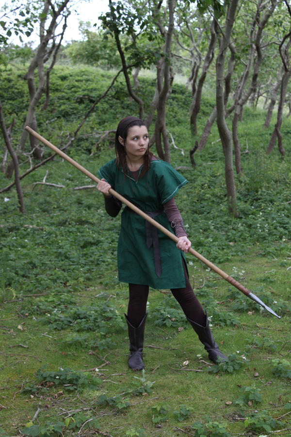 a woman dressed in an elf costume, complete with a green tunic and brown boots, stands in a wooded area holding a long spear.  she appears to be posing for a photograph, and her gaze is directed slightly to her right. the forest setting is lush and green, with numerous trees and branches visible. the overall mood is one of fantasy and adventure.
