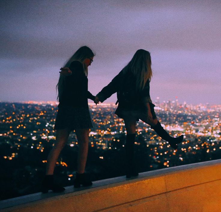 two girls stand on a ledge overlooking the city lights, silhouetted against the evening sky. they are holding hands and seem to be looking at the view.