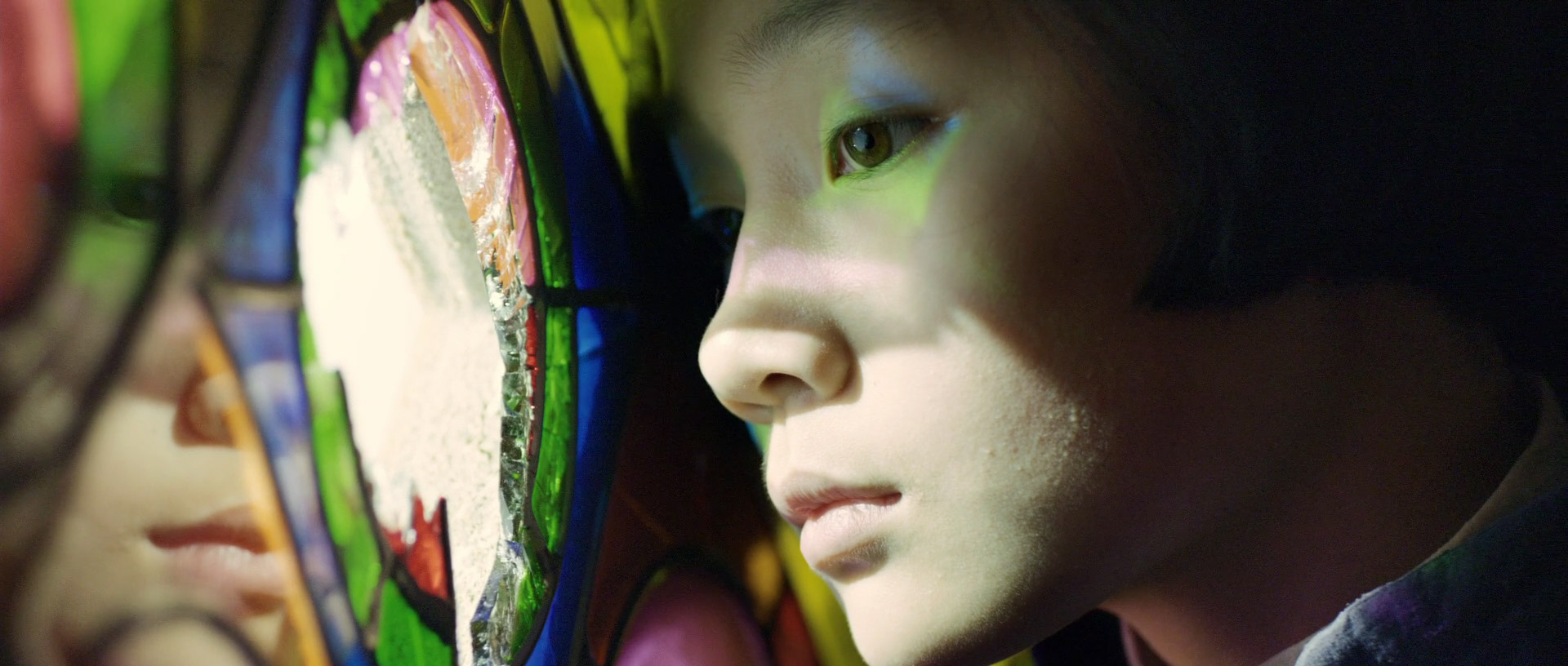 a closeup portrait of a young girl's face looking away from the camera.  she is standing in front of a stained glass window, and the colorful light is reflected in her face. the girl has a pensive expression on her face, and her eyes are downcast. the image is a study in light and shadow, and the girl's face is almost ethereal.  the stained glass is a beautiful and intricate design, with many different colors and shapes. the image captures the beauty and mystery of stained glass, as well as the innocence and sadness of childhood.