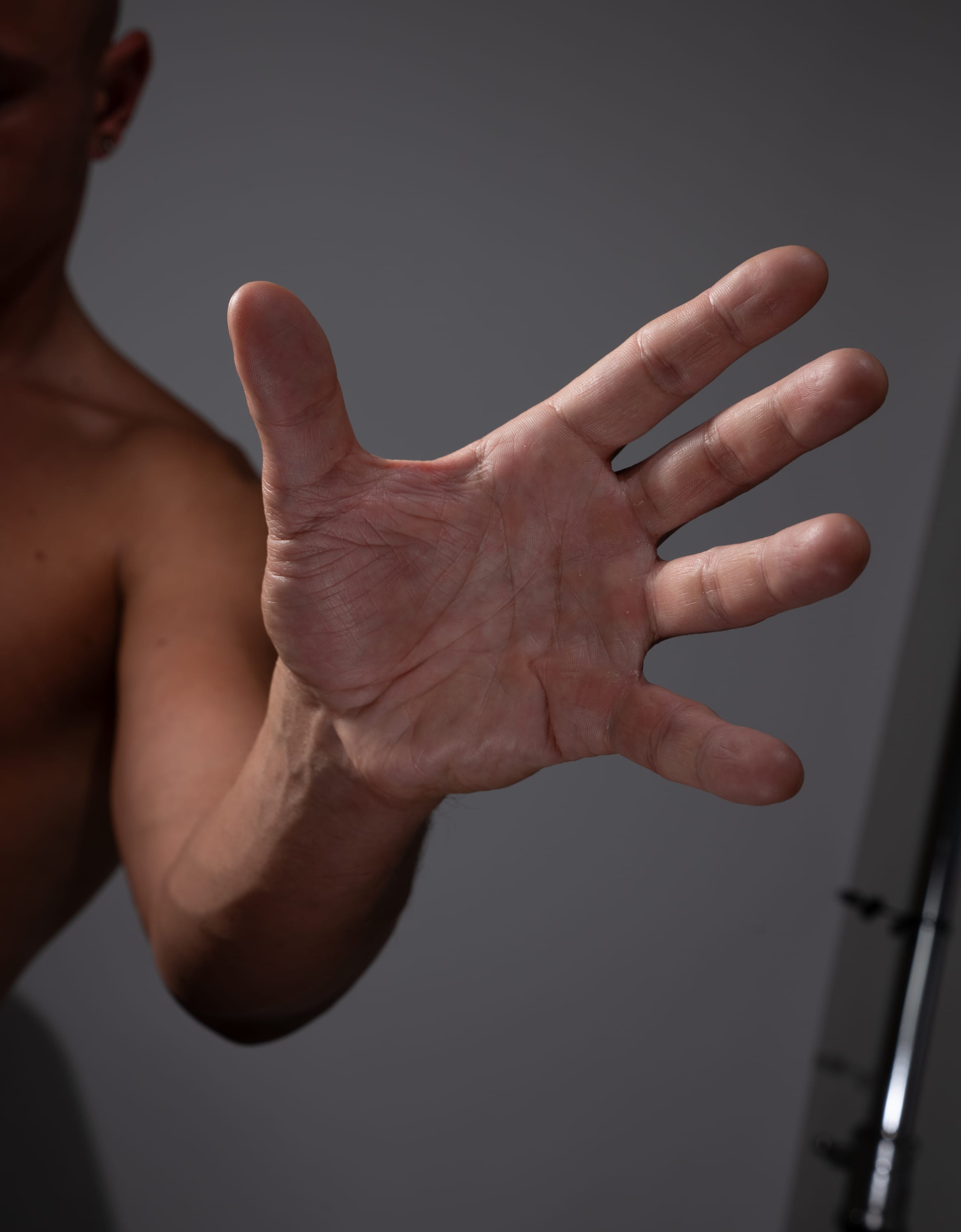 a closeup view of a man's outstretched hand against a neutral gray background. the hand is welllit, revealing the details of skin texture and muscle definition. the image focuses on the hand, with the rest of the man's body partially visible.