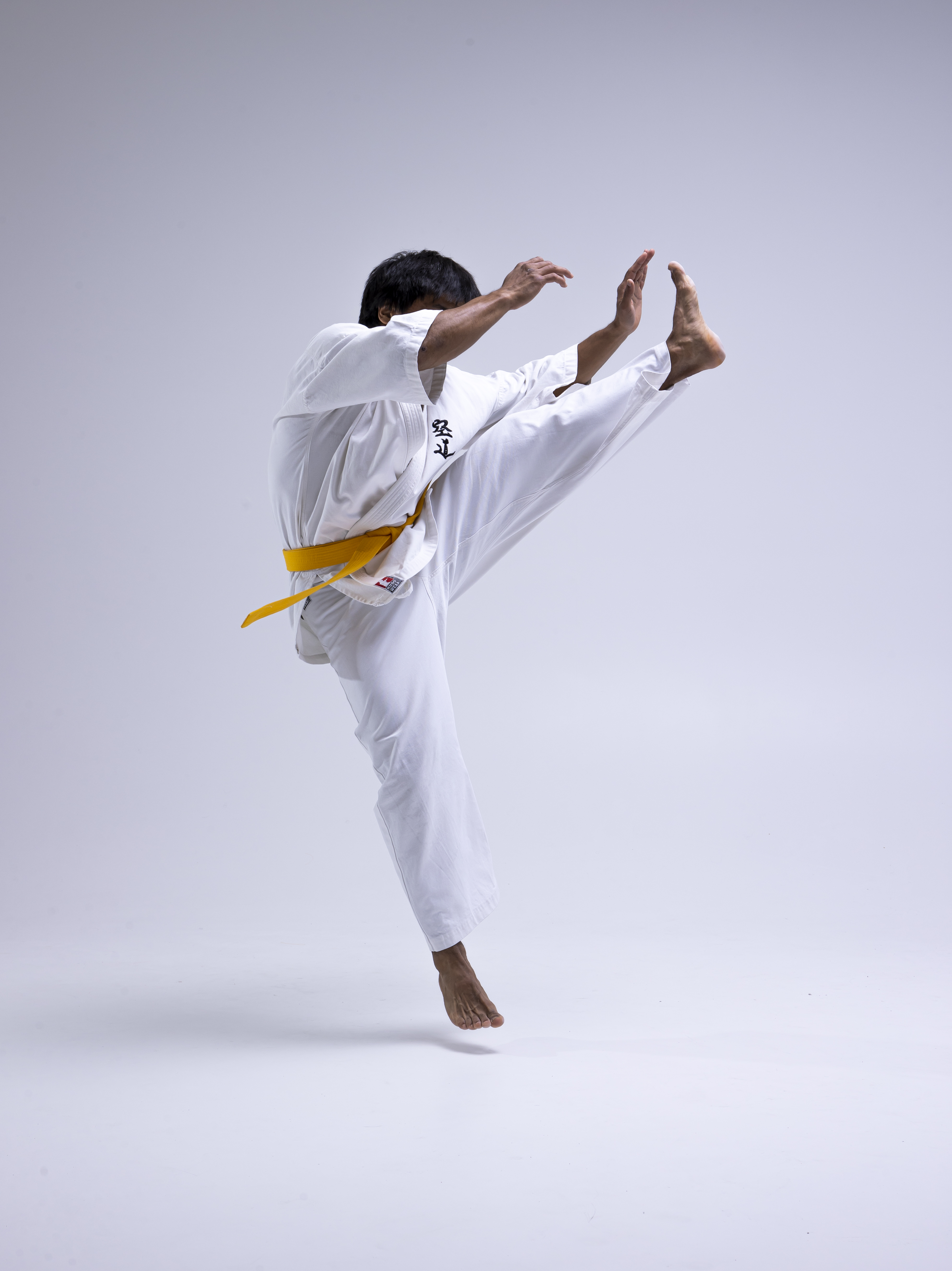 a person in a white karate uniform with a yellow belt is performing a high kick in a studio setting. the person is barefoot and suspended in midair, showcasing a dynamic athletic pose. the background is plain, emphasizing the action and movement of the martial artist.