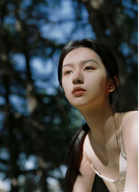 a young woman with dark hair tied back sits beneath a canopy of trees. the dappled sunlight filtering through the leaves casts a pattern of light and shadow on her face and surroundings. she is looking off into the distance with a contemplative expression. the overall mood is one of serenity and peacefulness.