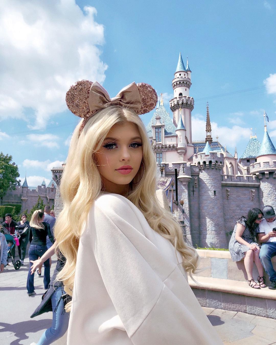 a photo of a young blonde woman posing in front of sleeping beauty's castle at disneyland. she is wearing mouse ears with a bow, and is wearing a sweatshirt. there are tourists in the background, the sky is blue and there are white clouds.