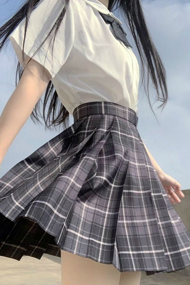 a woman in a school uniform with a white shortsleeve shirt and a plaid pleated skirt. the skirt is black and grey with white lines. she is standing outside against a blue sky. her hair is long and black.