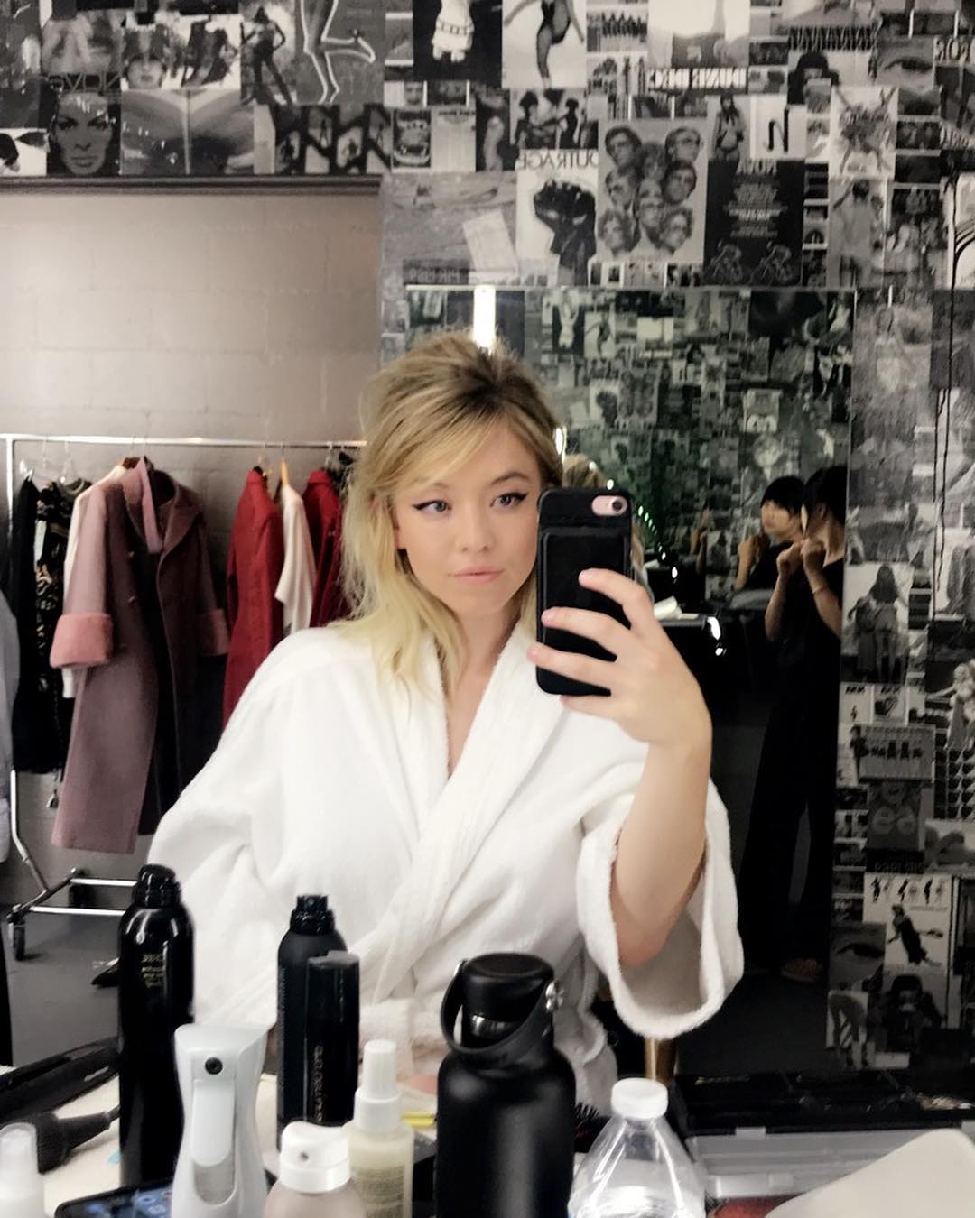 the image shows a young blonde actress, likely backstage at a photoshoot or event. she's wearing a white bathrobe and is taking a selfie in a mirror. the mirror reflects a room with clothing racks displaying various coats and outfits, along with hair and makeup products. the background features a striking wall covered in a collage of black and white photographs. another woman can be seen in the background, likely a stylist or member of her team. the overall impression is one of a behindthescenes moment during a fashion or media event.