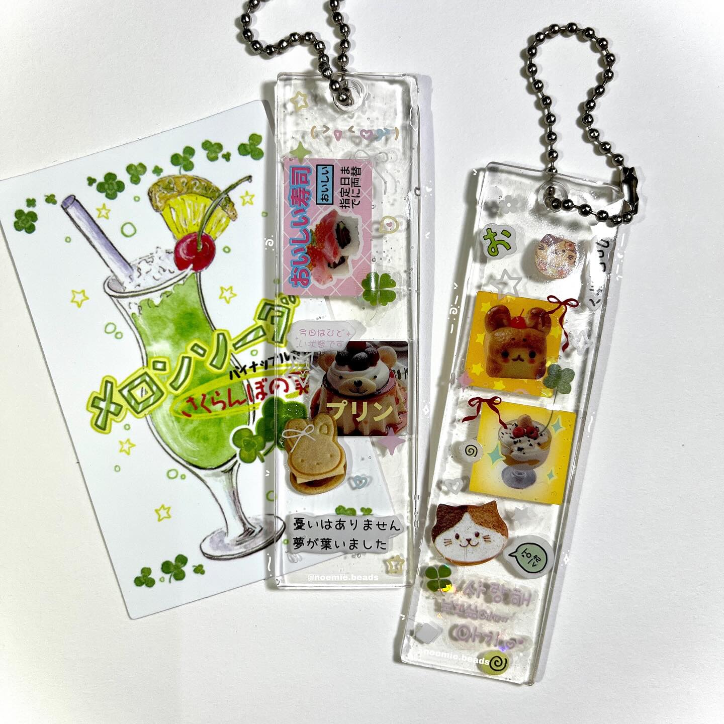 this image showcases a variety of charming trinkets and accessories. there's a card featuring a melon soda drink adorned with a pineapple wedge and cherry, surrounded by lucky clovers and japanese text. additionally, two clear charms are present, each filled with an array of delightful food items like sushi, pudding, and sandwich cookies, along with cartoon characters and more japanese text. these charms are complemented by a beaded chain, enhancing their appeal.