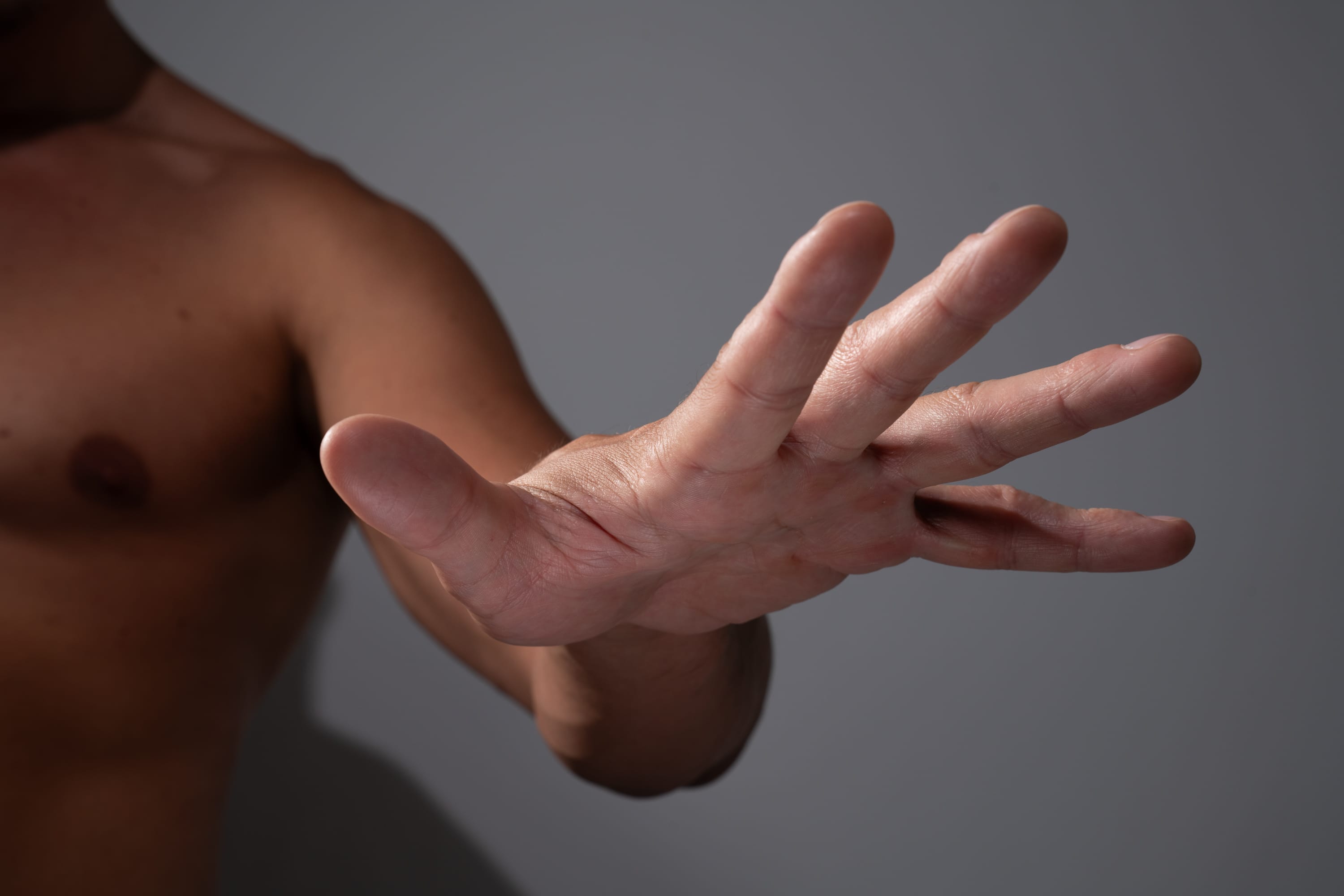 a shirtless male extends his open right hand, fingers spread, toward the viewer in a gesture of halting or signaling "stop." the focus is on the hand and arm, with the man's torso blurred in the background. the lighting is soft and even, creating a sense of calm and control.