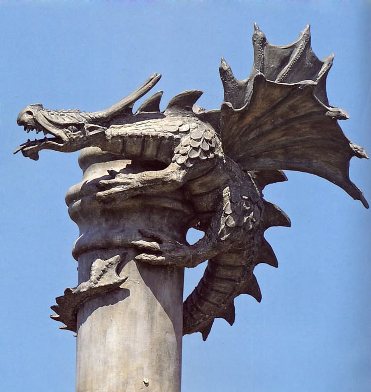 the image shows a detailed stone or bronze sculpture of a dragon perched atop a fluted column against a clear blue sky. the dragon is depicted with intricate scales, sharp claws gripping the column, and large, ornate wings. the style suggests a medieval or gothic influence.