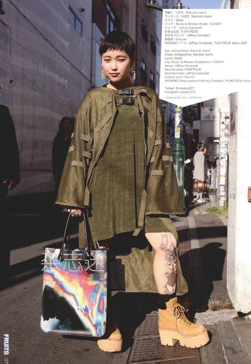a young woman poses on a city street, showcasing a unique and edgy fashion sense. she wears an olive green, belted tunic dress, platform combat boots, and a large tote bag with an abstract, iridescent design. her short hair and arm tattoo add to the overall edgy aesthetic. the image appears to be taken from a japanese street fashion magazine, as indicated by the japanese text alongside the photo.