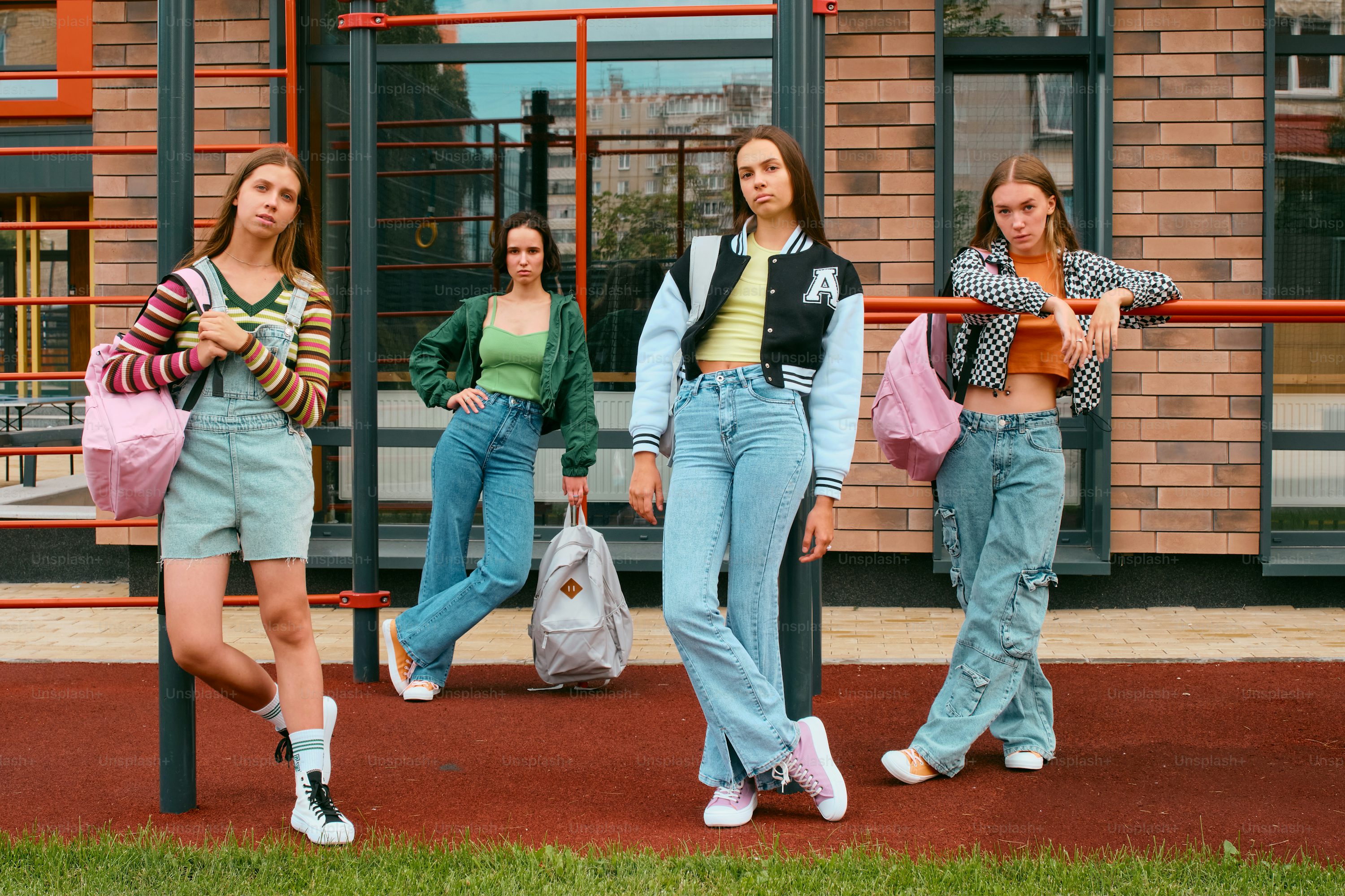 four teenage girls are standing in front of a brick school building. they are all wearing casual clothes, including jeans and backpacks. some are leaning on a railing, and appear to be happy and confident. the overall atmosphere is one of friendship and carefree youth.  the scene is brightly lit, suggesting daytime.