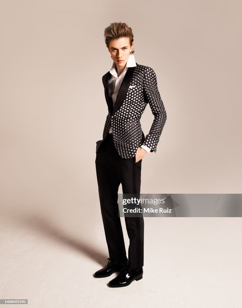 a full length studio shot of a man in a fashionable suit. he is standing with one hand in his pocket and a serious expression on his face. he is wearing a black and white polka dot jacket, a white shirt, black pants, and black shoes.