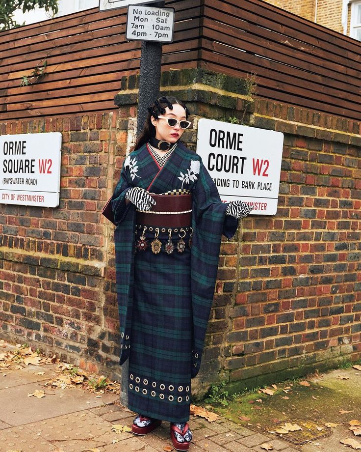 a woman is standing on a street corner next to brick walls and street signs. she is wearing a dark blue and green plaid kimono with white floral accents, a wide brown belt, striped gloves, and sunglasses. her footwear is maroon colored. there is a street sign that says "orme square w2 (bayswater road) city of westminster" and another sign that says "orme court w2 ading to bark place westminster". a third sign indicates "no loading mon  sat 7am  10am 4pm  7pm".