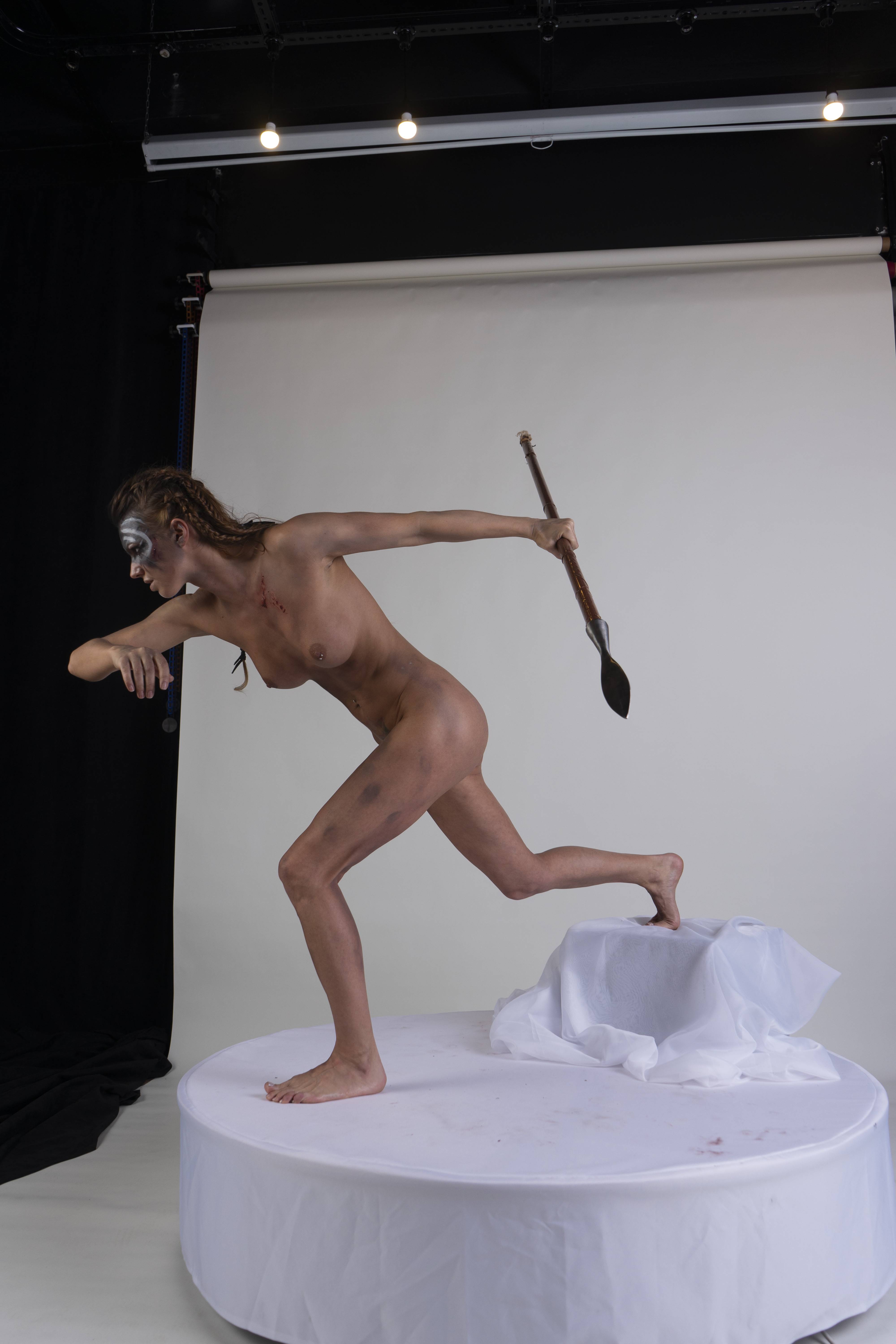a nude female warrior with body paint on her face and bruises on her body runs on a white platform in a studio setting. she holds a spear in her right hand, her body angled in profile against a white backdrop.