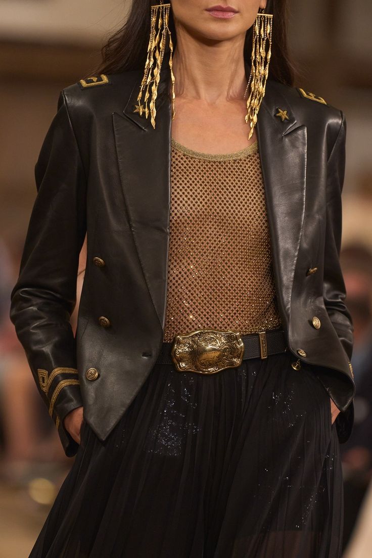 a woman wearing a black leather jacket with gold accents over a sheer, sparkly gold top. she is also wearing a gold belt with a large buckle, and long gold earrings. her hair is styled in a sleek, straight style, and she is looking off to the side. she is dressed in an elegant and glamorous fashion.