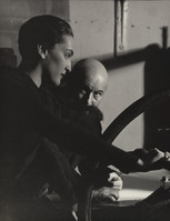 the black and white photograph depicts a closeup view of a man and woman seated close together, appearing intimate. the woman, younger, is positioned prominently in the foreground, her face partially obscured by shadow, while the older man sits somewhat behind and to the side. the setting appears to be a dark, industrial workshop or studio, suggested by the visible presence of a large, potentially mechanical wheel in the background.  the lighting is dramatic and uses strong contrasts between light and shadow, contributing to a moody and evocative atmosphere. the overall style is reminiscent of film noir photography of the 1930s or 1940s.  the focus is primarily on the emotional connection between the couple, as they seem to be close together and intimate.