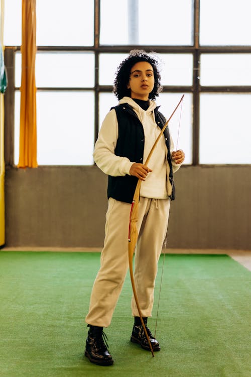 a young woman stands in an indoor archery range, holding a wooden longbow. she is wearing a beige sweatshirt, black vest, and black boots. the background is a bright room with large windows. the young woman looks directly at the camera with a determined expression.