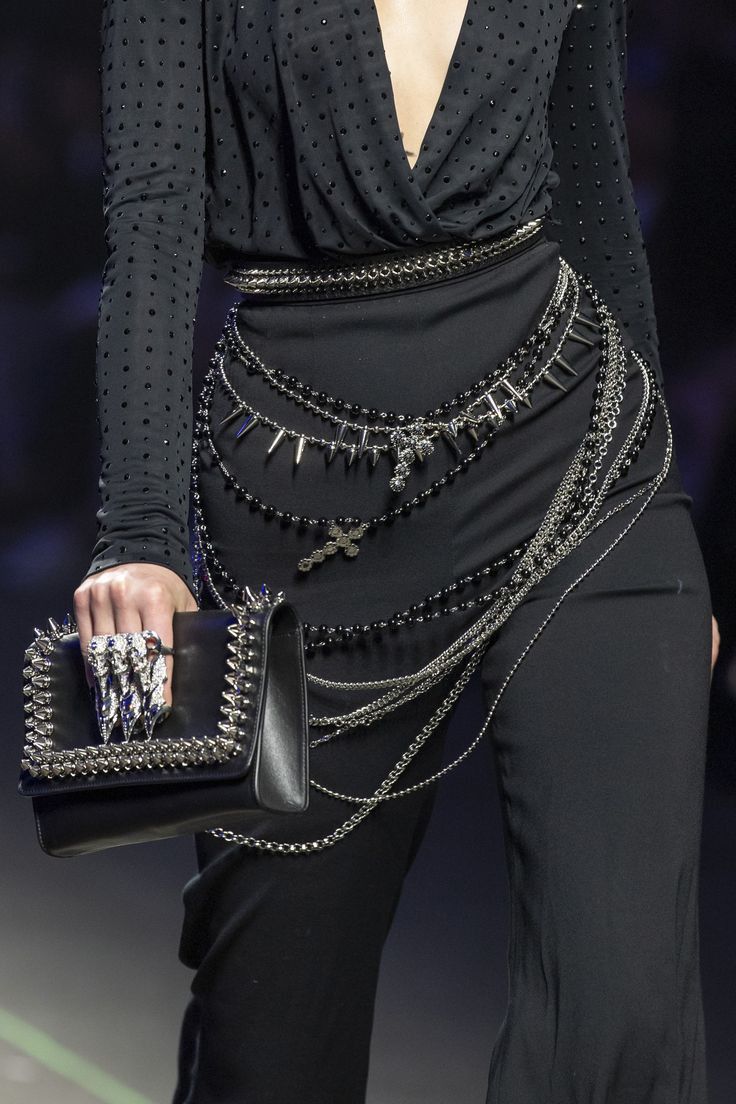 a model wearing a black dress with long sleeves and a plunging neckline.  she is wearing several silver chains and a black choker around her neck. the model is also wearing a black clutch bag and black pants.