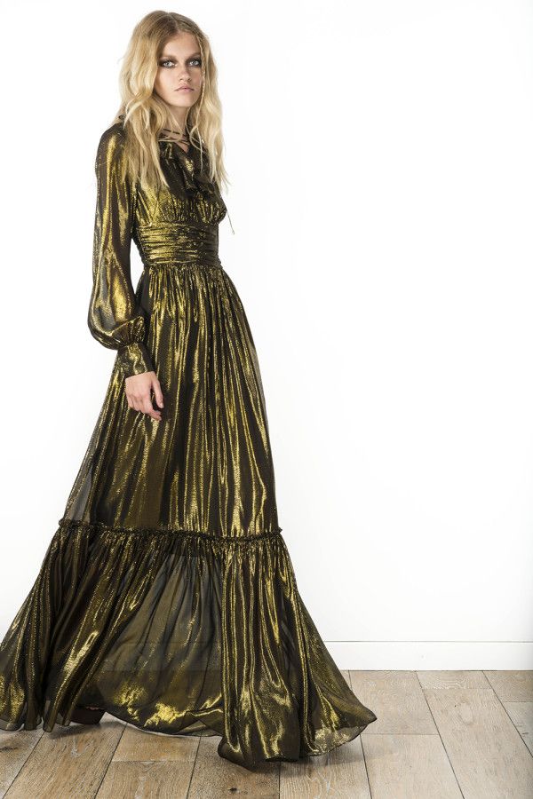 a fulllength image of a blonde model wearing a long, flowing gold metallic dress. the dress has long sleeves, a pleated bodice, and a ruffled hem. the model is standing on a wooden floor against a white background. the dress appears to be made of a sheer or semisheer fabric that shimmers in the light. the overall style is elegant and romantic.