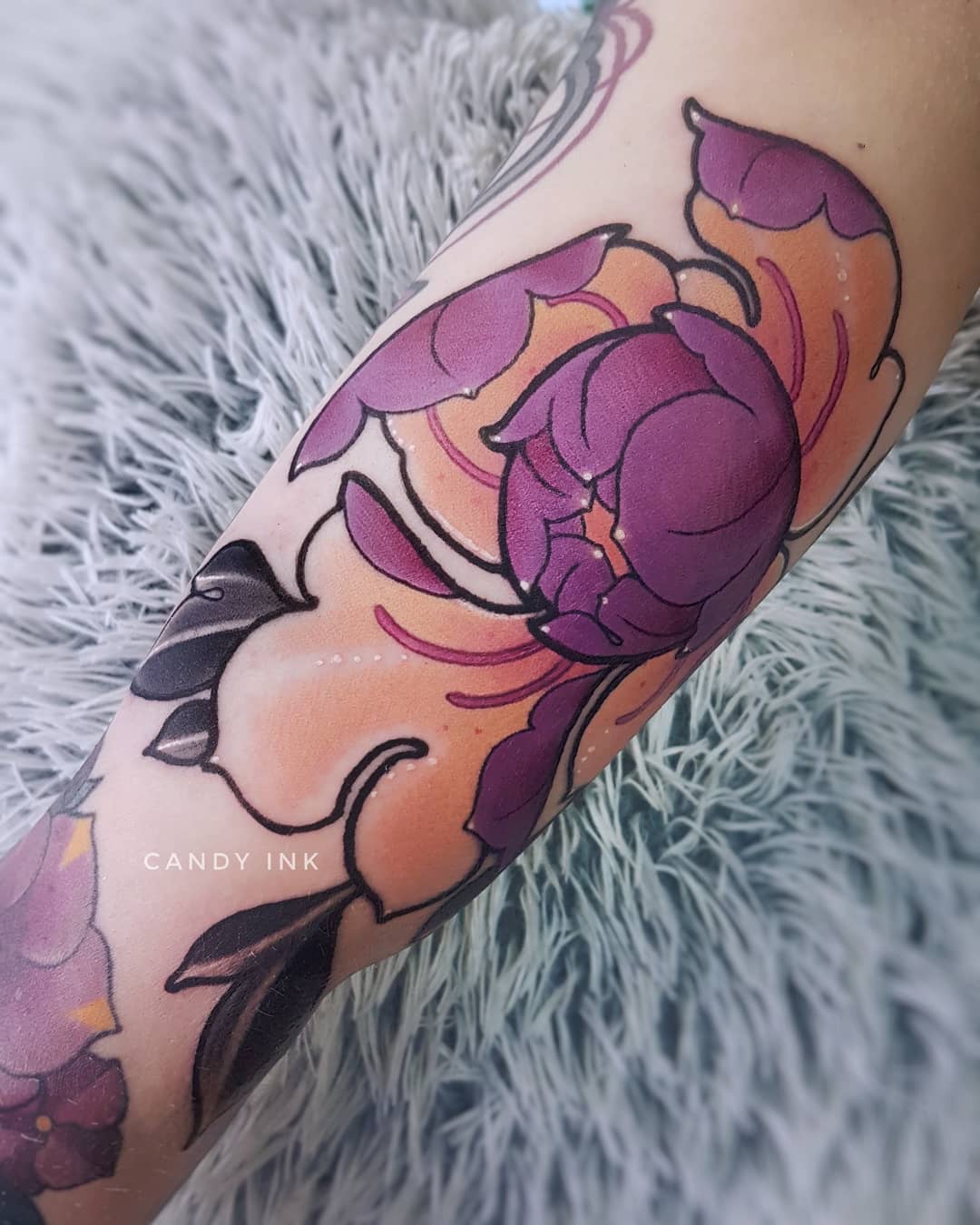 a closeup photograph showcasing a neotraditional style tattoo of a peony flower on a person's forearm. the tattoo features shades of purple, orange, and black ink. the background reveals a glimpse of a fuzzy, creamcolored surface.
