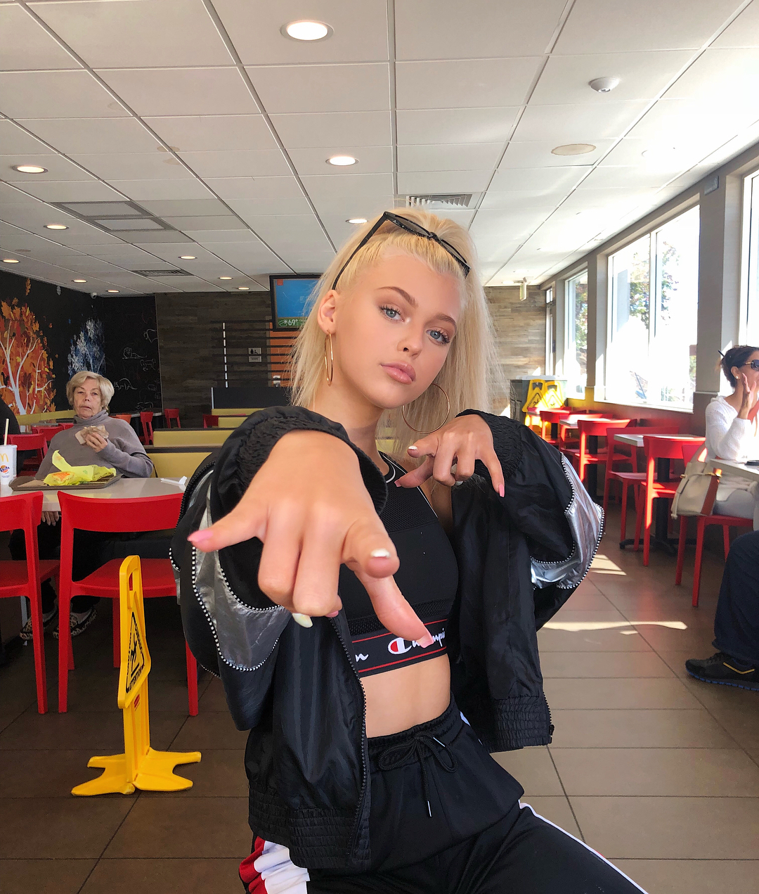 a young woman with blonde hair and sunglasses on her head, wearing a black crop top, black jacket, and black sweatpants, is sitting at a table in a restaurant and pointing towards the camera. there is a yellow caution sign near her and other patrons in the background.