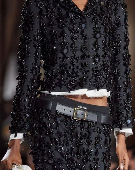 a woman wearing a black, beaded dress, jacket, and belt on a runway. she is wearing a highfashion outfit that is both elegant and chic. the outfit is full of embellishments, sequins, and beading. she is standing on a runway.  the model is posing for the camera.